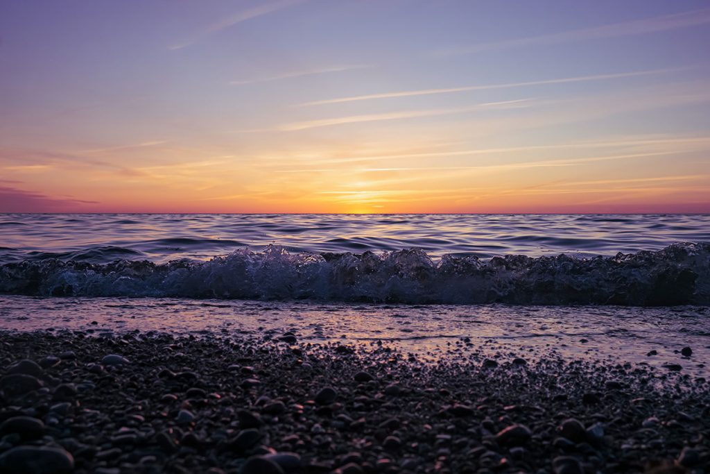 Fotografie eines lila Sonnenuntergangs an der Ostsee mit Kiesstrand im Bokeh und sanften Wellen.